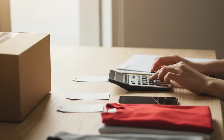 Mãos usando uma calculadora na bancada, ao lado de etiquetas de preço e peças de roupa dobradas